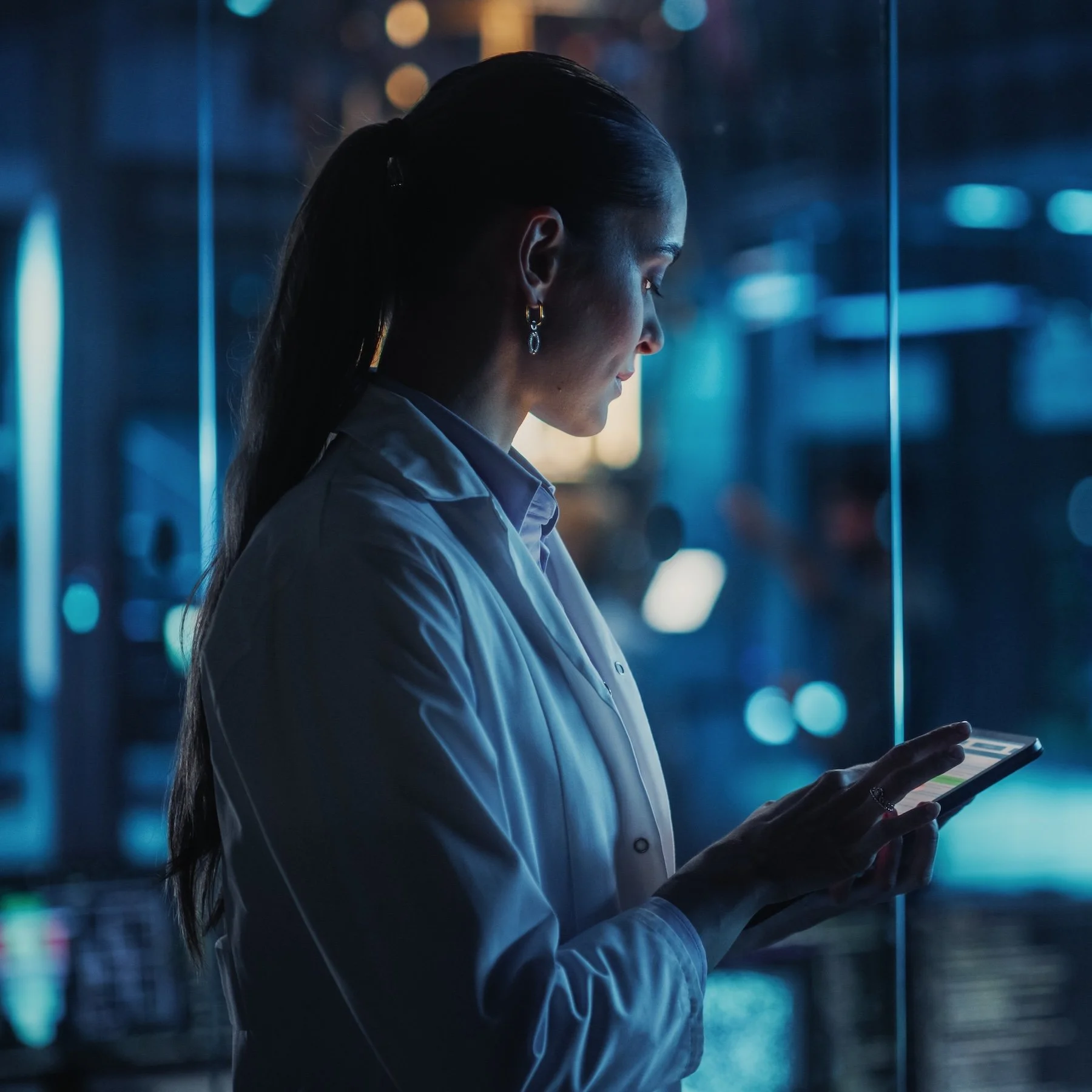 A woman in a white lab coat with dark hair tied back, wearing earrings, looking at her smartphone in a modern urban setting at night with blurred city lights in the background.