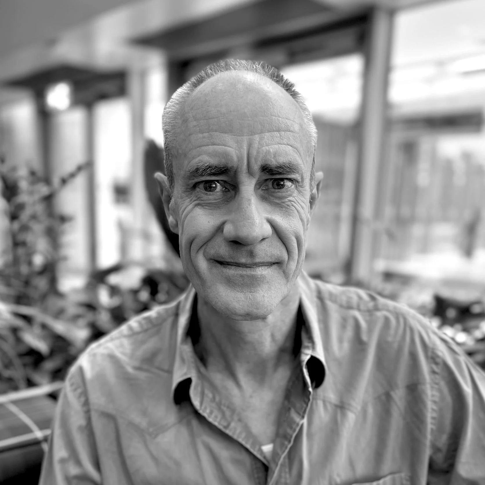 A close-up portrait of an elderly man with a bald head, smiling and wearing a collared shirt, in a blurred background of what appears to be a greenhouse or botanical setting.
