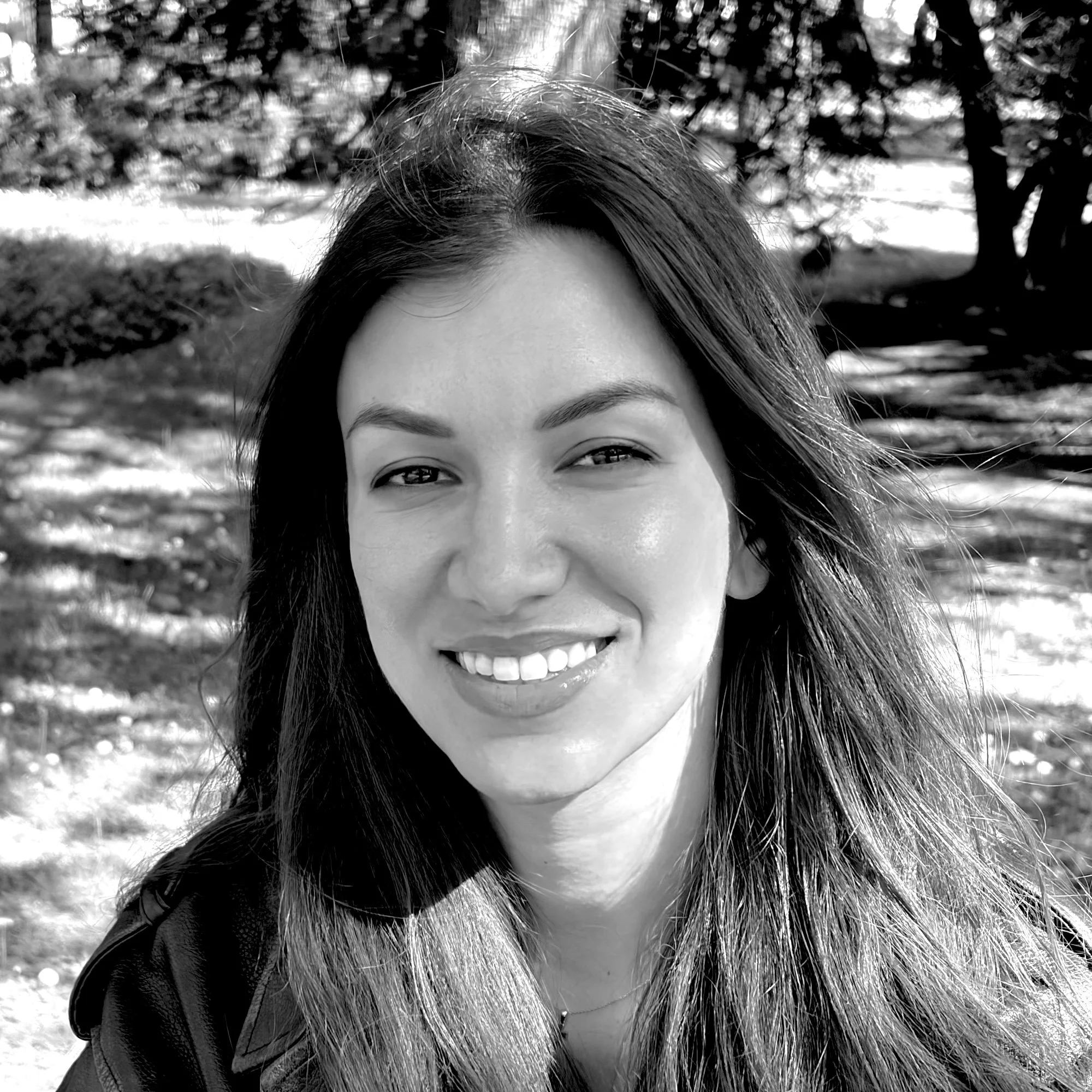 Black and white photograph of a woman smiling outdoors with trees and grass in the background.