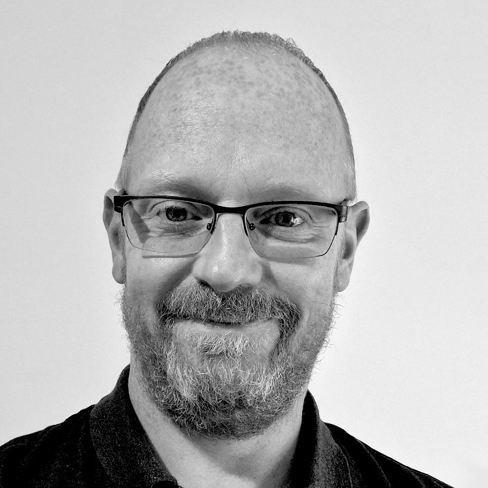 Black and white photo of a smiling man with glasses, a beard, and short hair, wearing a dark shirt, against a plain background.