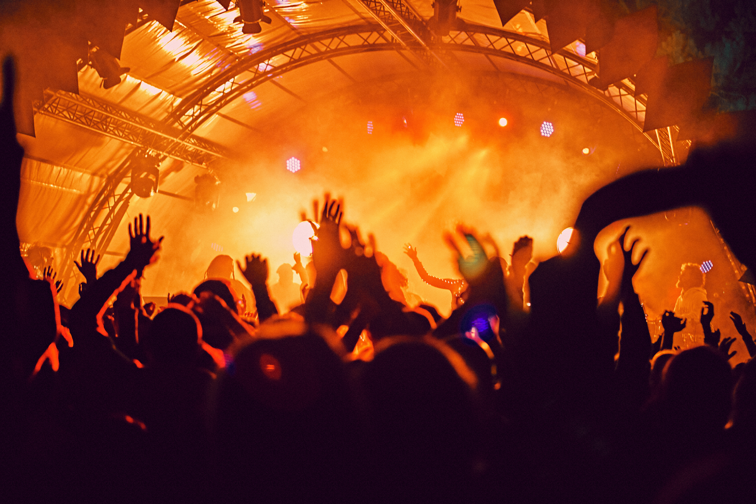 A crowd of people dancing and raising their hands at a concert or music festival with a brightly lit stage and orange lighting.