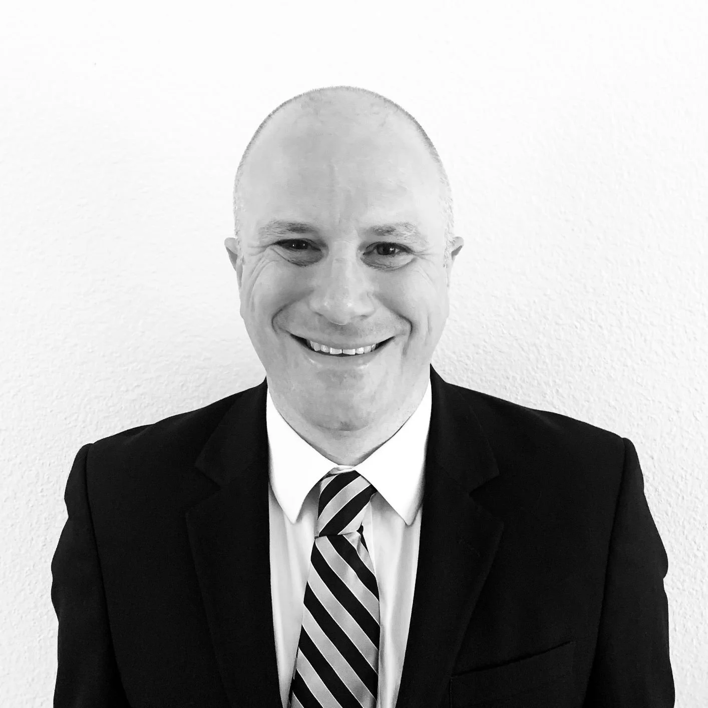 A smiling man with a shaved head wearing a black suit, a white shirt, and a striped tie, standing against a plain white wall.