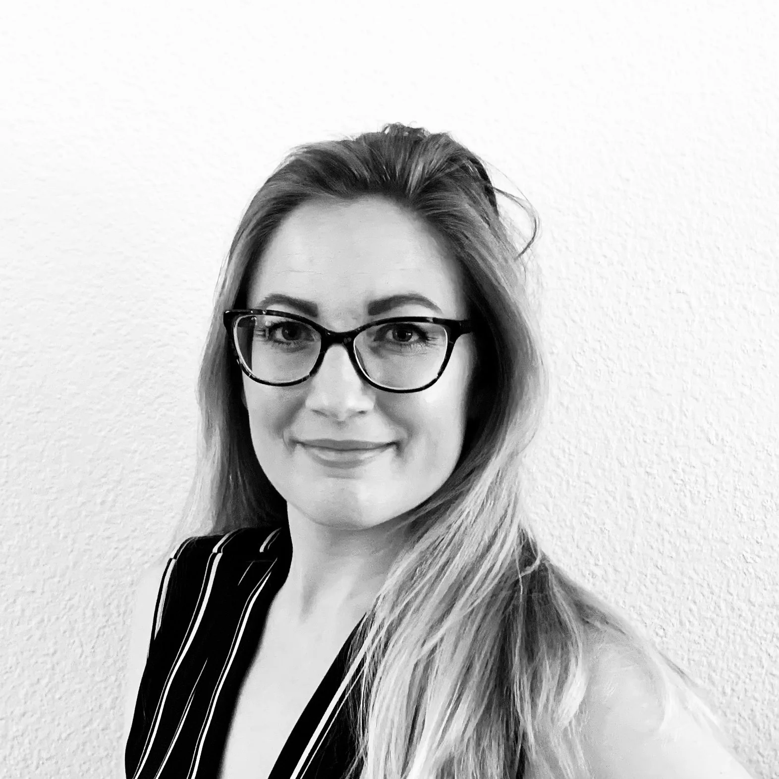 Black and white portrait of a woman with glasses, long hair, wearing a pinstriped shirt, smiling slightly, standing against a plain wall.