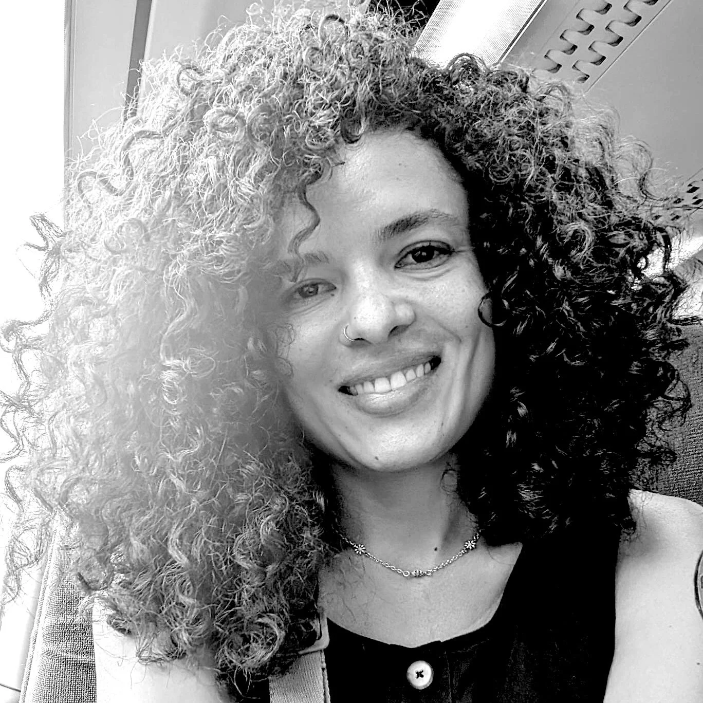 A woman with curly hair smiling, wearing a nose ring, necklace, and a buttoned top, standing indoors.