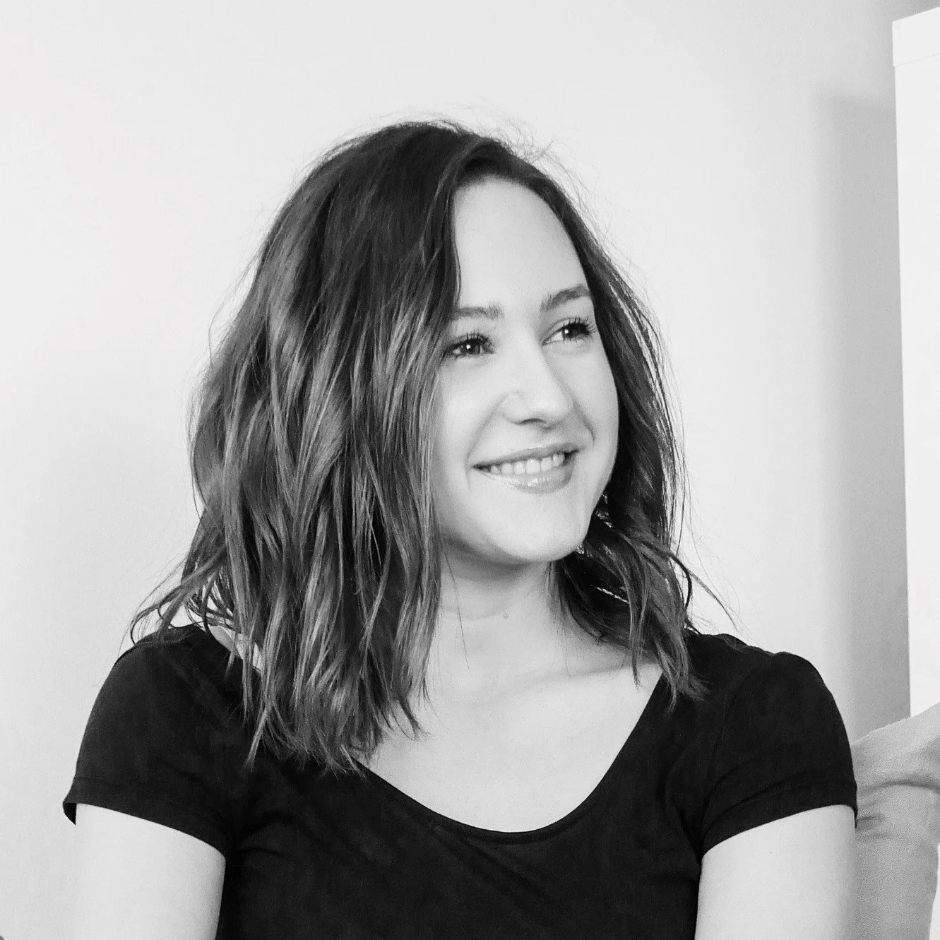 A smiling woman with wavy, shoulder-length hair wearing a black shirt, sitting in front of a plain wall.