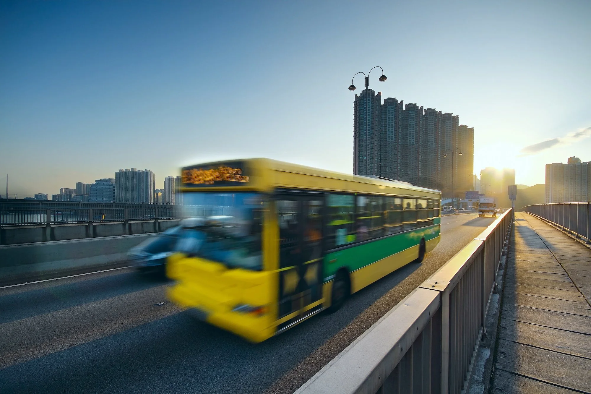 A city highway during sunset with a green and yellow bus moving quickly in the foreground, and tall residential buildings in the background. The sky is clear with a few clouds.