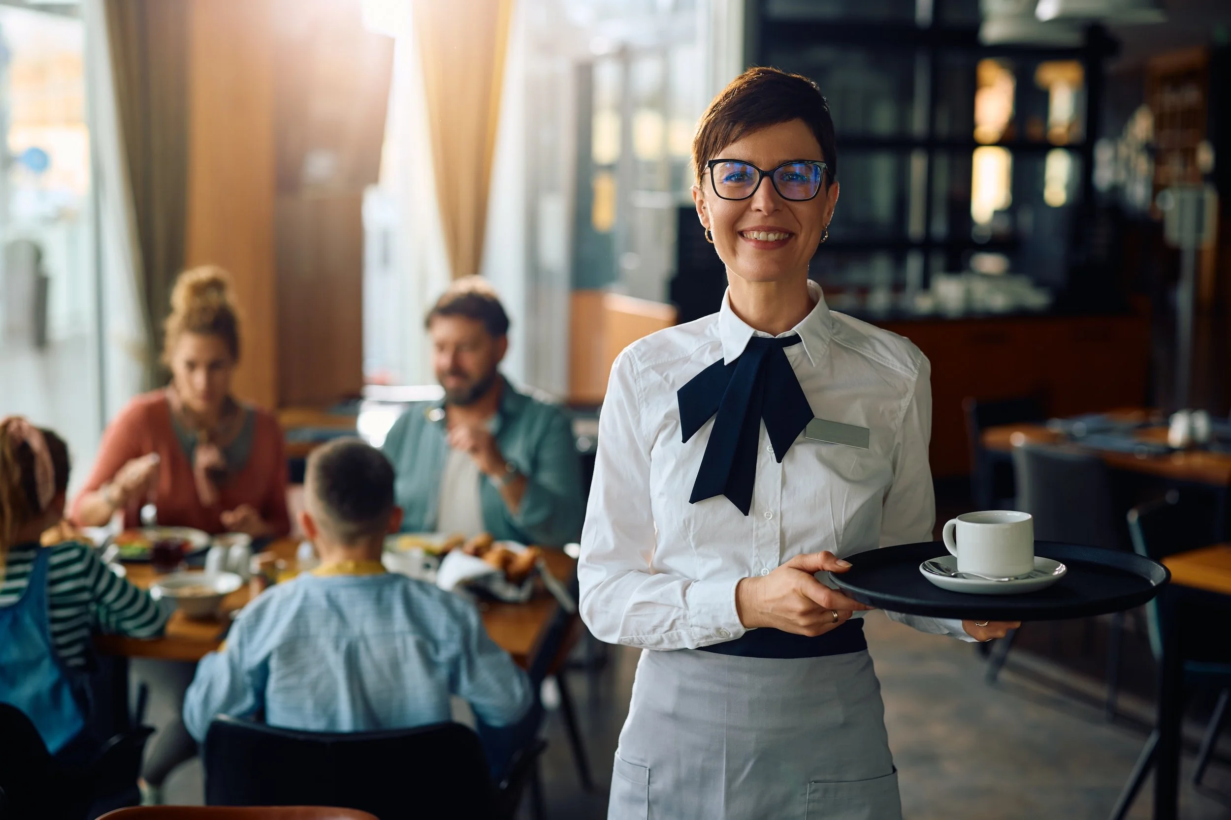A smiling female waiter with short dark hair and glasses, dressed in a white shirt with a black bow tie, serving coffee or tea on a tray in a busy restaurant. In the background, there are customers, including children and adults, dining and socializing.