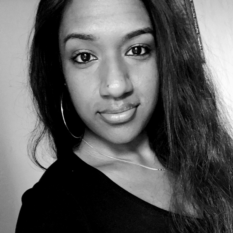 A close-up black and white photo of a young woman with long dark hair, wearing earrings, a necklace, and a black top, smiling subtly.