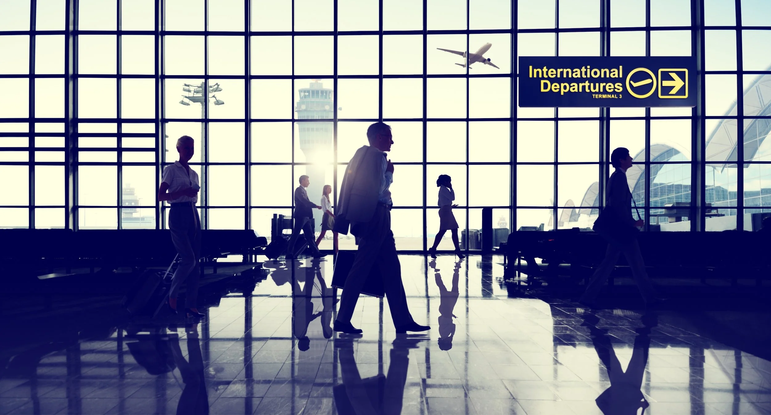 Silhouettes of travelers walking through an airport terminal with large windows, with airplanes visible outside and a sign pointing to international departures.