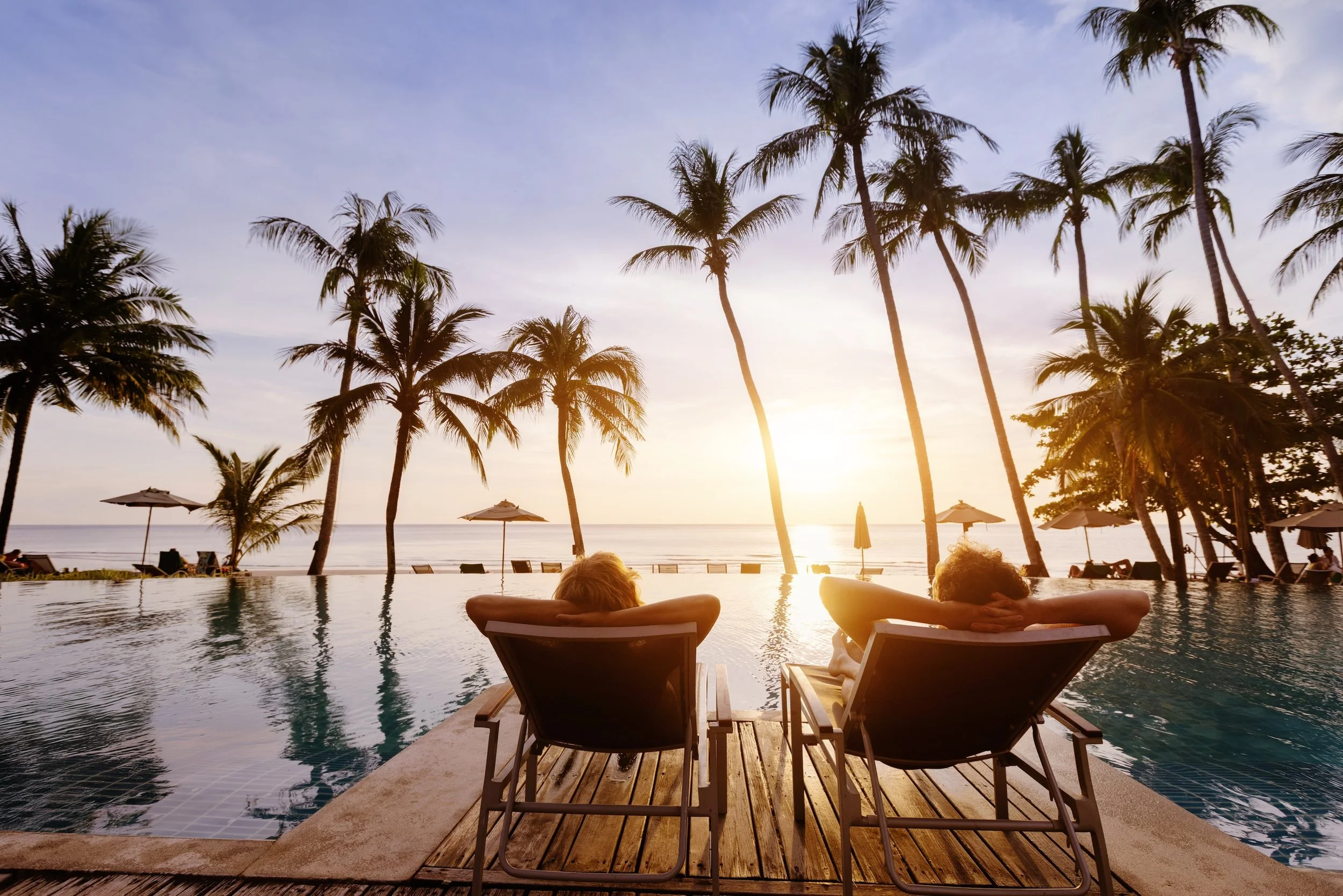 Two people relaxing in lounge chairs by a swimming pool at sunset, with palm trees and umbrellas in the background.