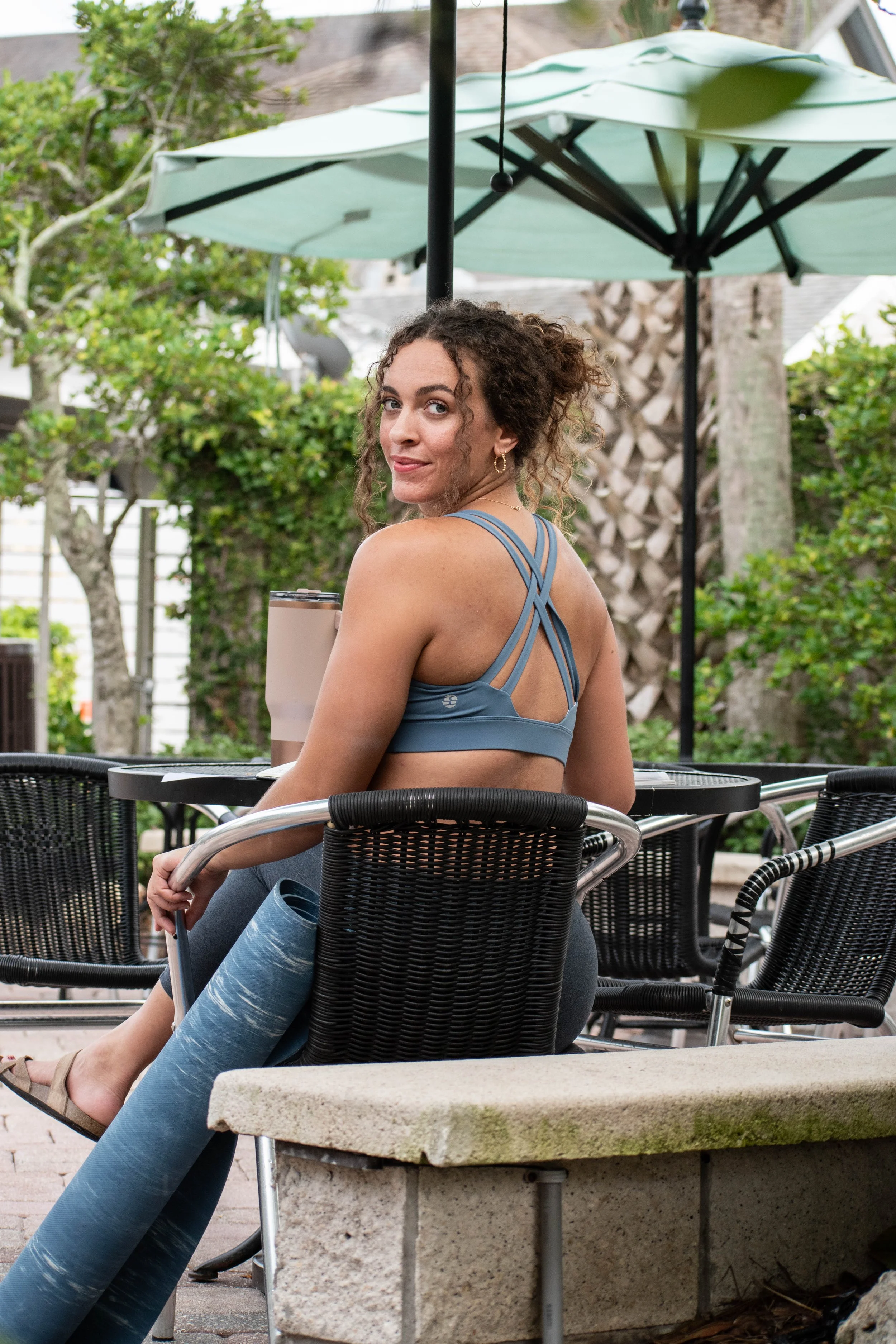 private yoga teeacher sitting outside in a courtyard at a table with an umbrella. she is looking over her shoulder directly into the camera