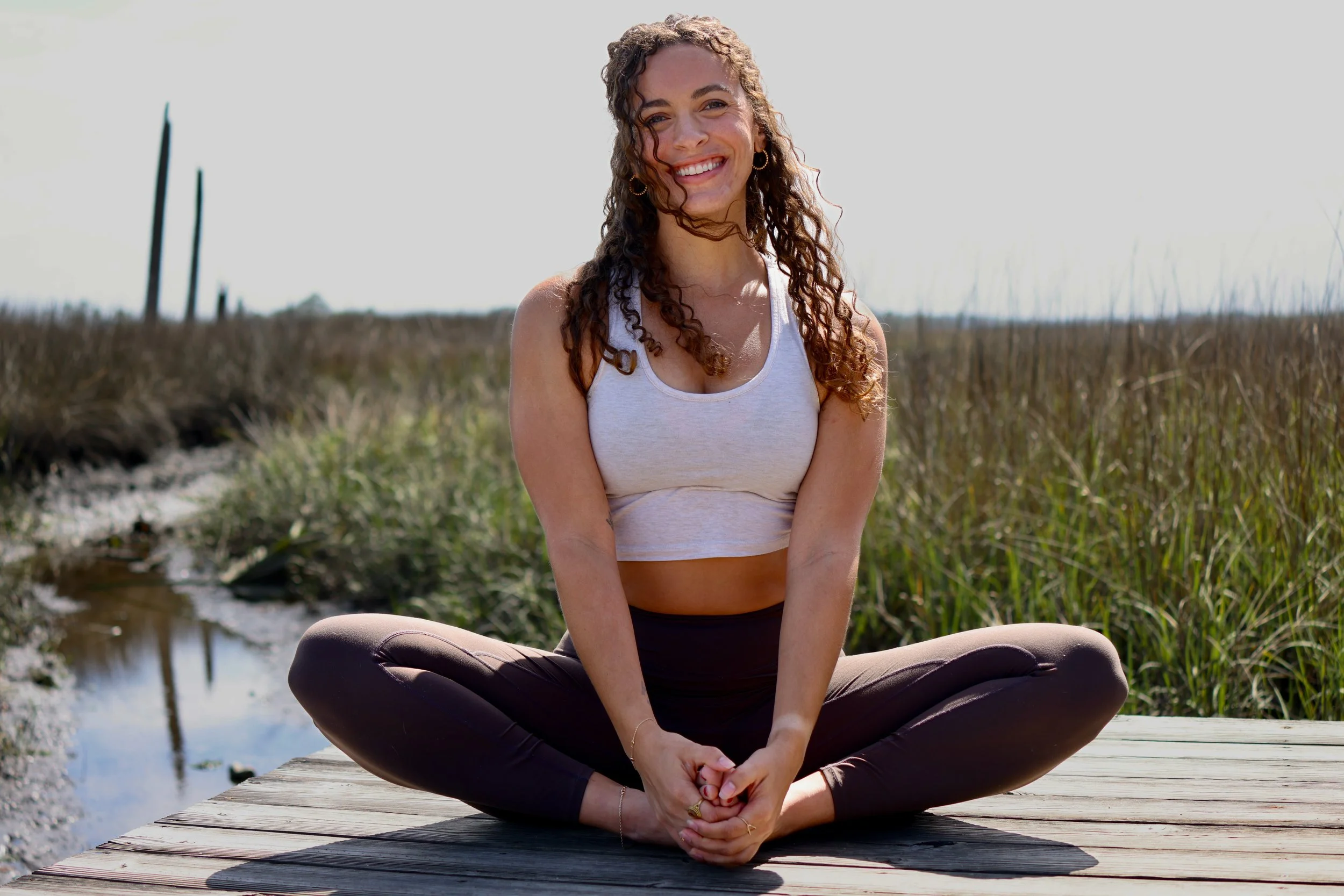 private Yoga teacher in a seated butterfly position. she is looking at the camera and smiling. the wind is blowing her hair across her face