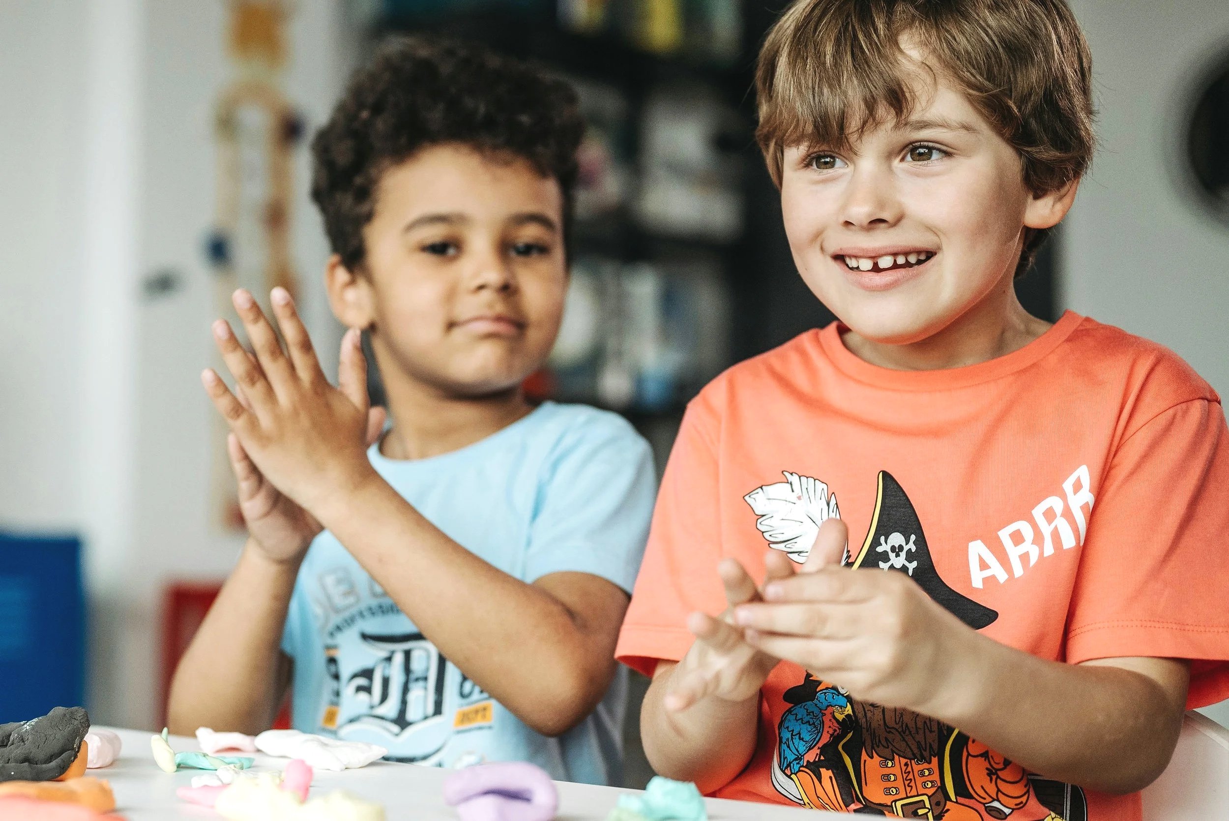 Two young boys sitting at a table, engaging with colorful playdough food. The boy on the left has curly hair and a light blue shirt, while the boy on the right has straight hair and is smiling, wearing an orange shirt with a pirate graphic.