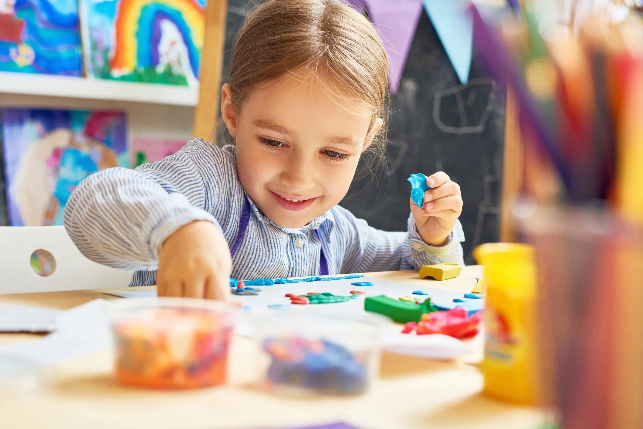 A young girl with brown hair, wearing a striped shirt, is smiling and playing with colorful modeling clay at a table.