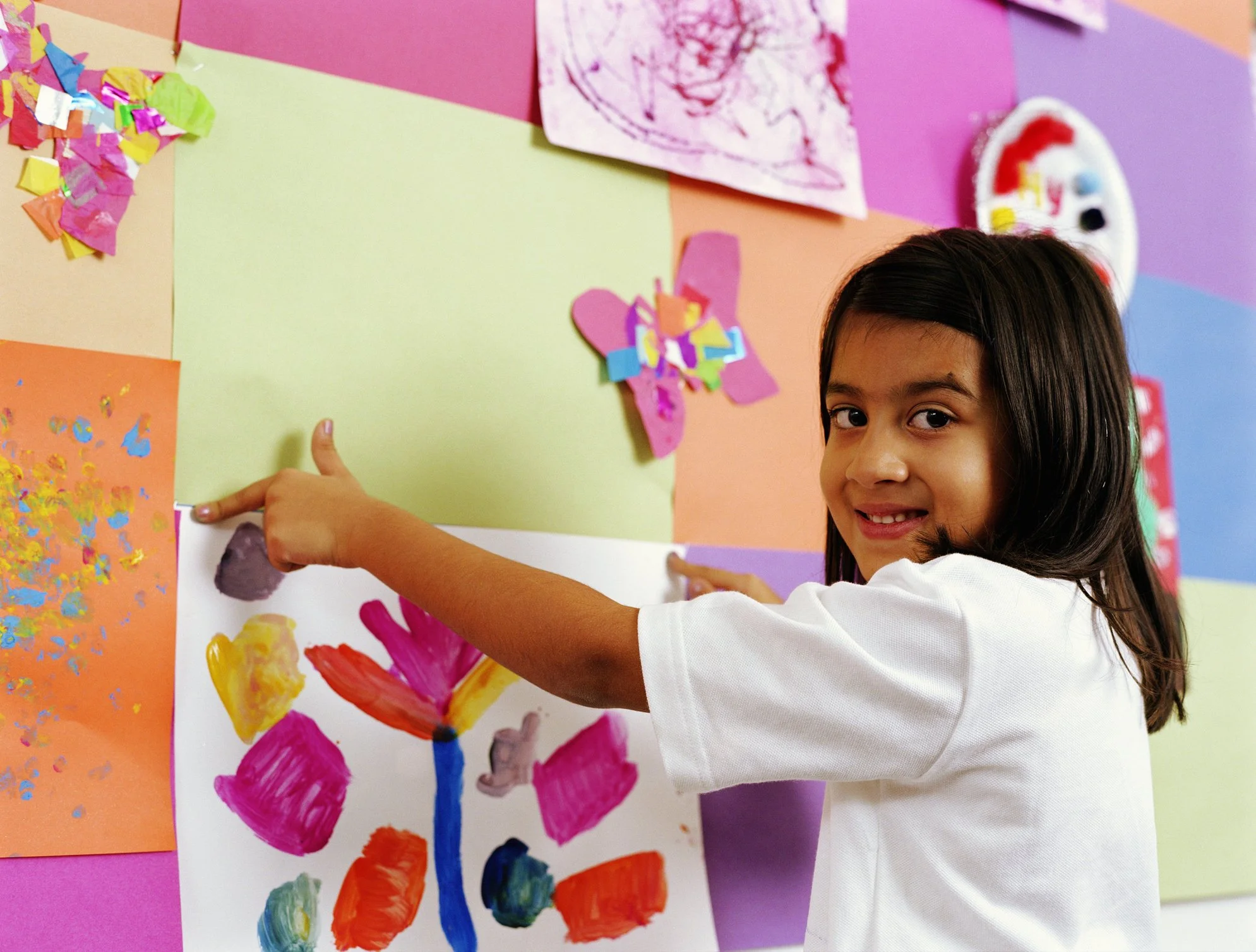 A young girl in a white t-shirt points to her colorful painted flower on a bulletin board decorated with artwork, on a wall painted in pastel colors.
