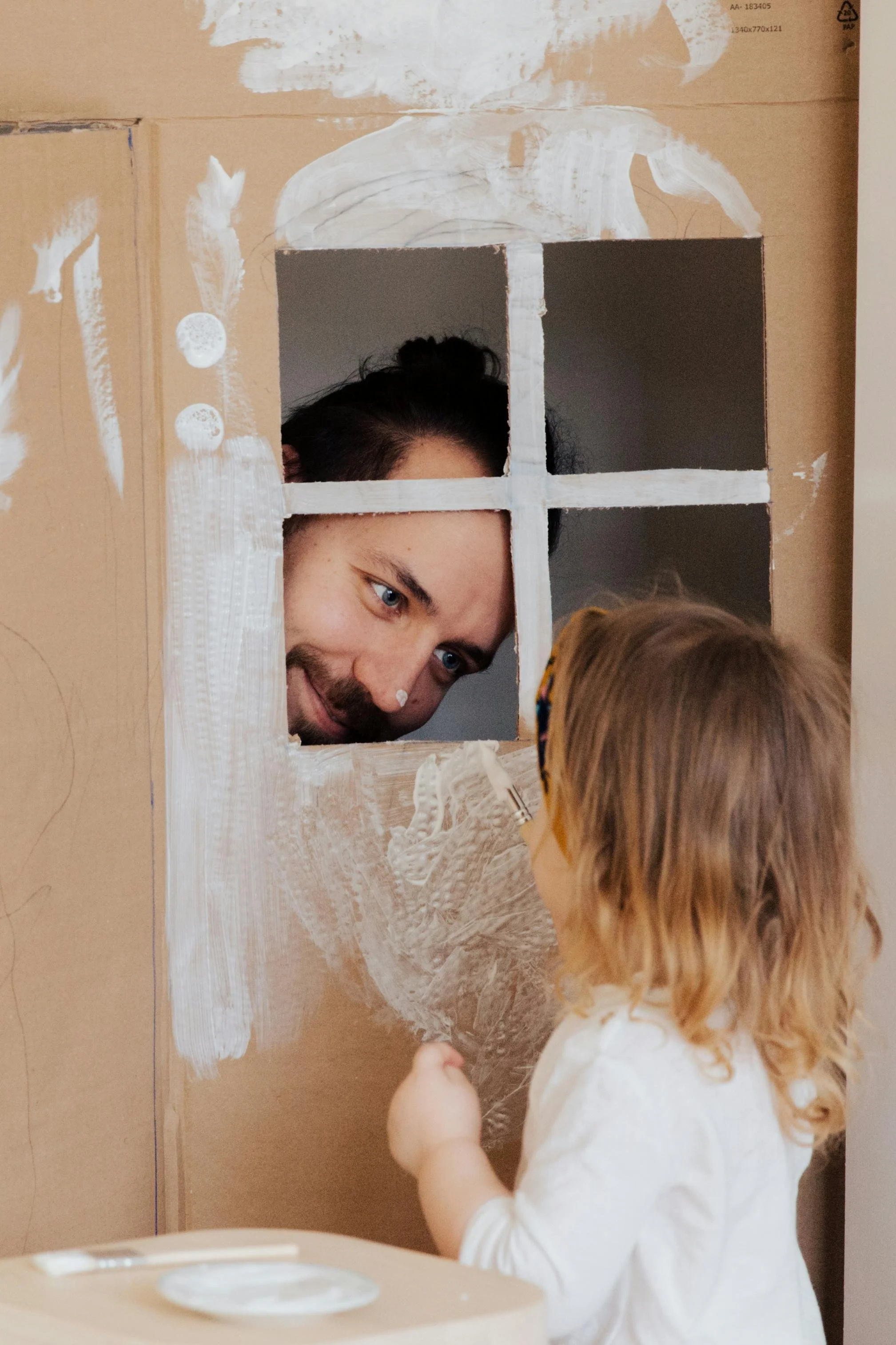 A man is peering through a painted window opening in a cardboard box, engaging with a young girl who is holding a paintbrush. The scene depicts a playful, creative activity involving painting and pretend play.
