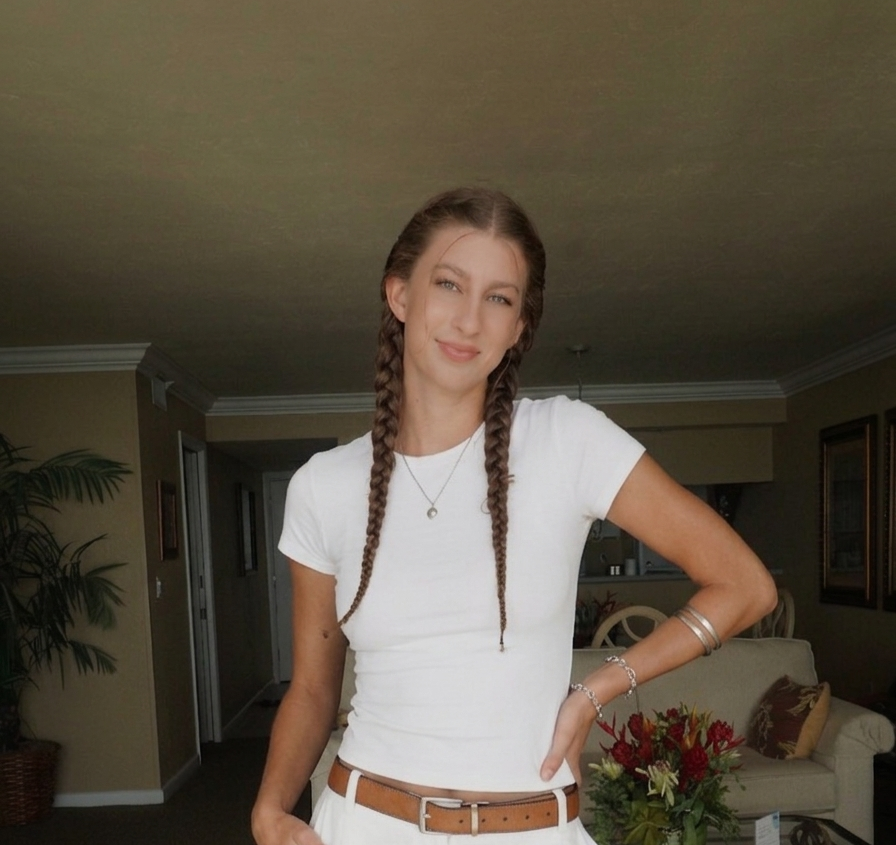 A young woman with braided hair, wearing a white t-shirt and light-colored pants, standing in a living room with a neutral expression.