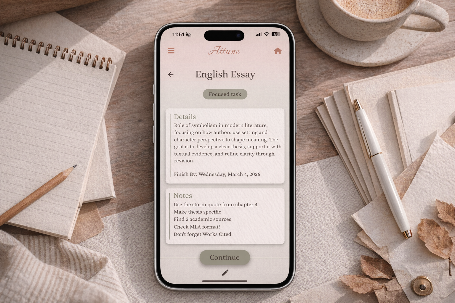 A smartphone displaying a writing app for an English essay with notes and details, surrounded by notebooks, a pen, a cup of coffee, and decorative paper and dried leaves on a wooden desk.
