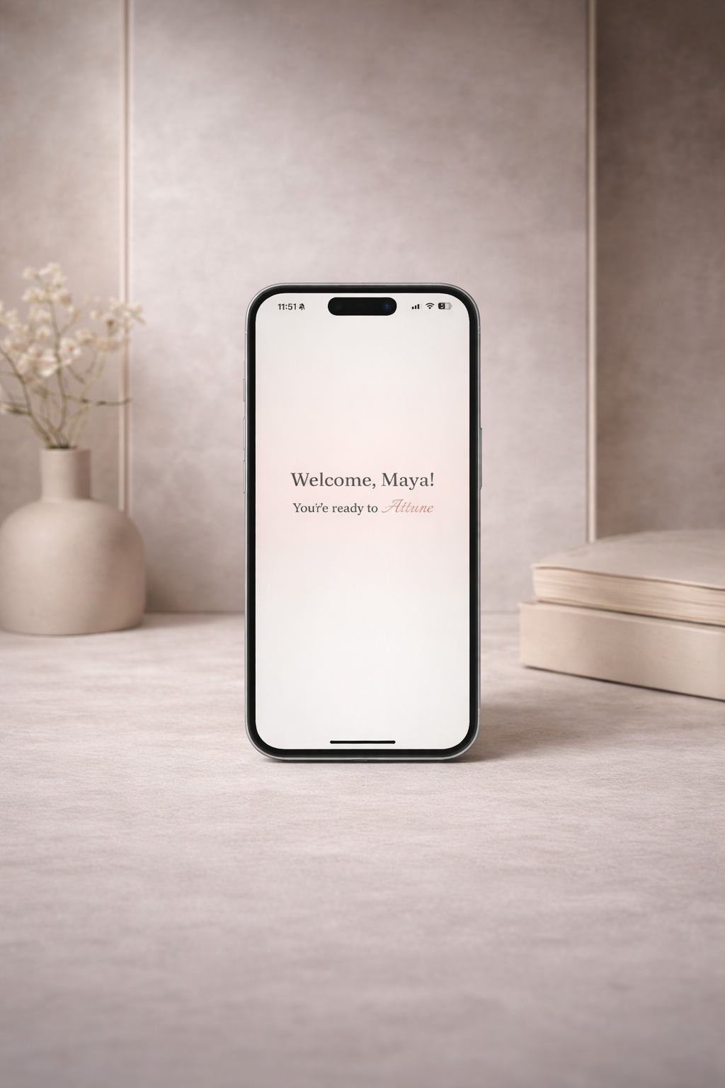 A smartphone displays a welcome screen with the message "Welcome, Maya! You're ready to Attune" on a beige table with a vase of white flowers and a closed book in the background.