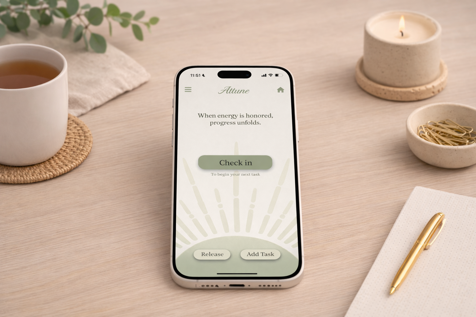 Smartphone on a wooden desk displaying a wellness app called Attune with a check-in button. The desk also has a mug of tea on a woven coaster, a lit candle, a bowl with gold paper clips, a white notepad, and a gold pen.