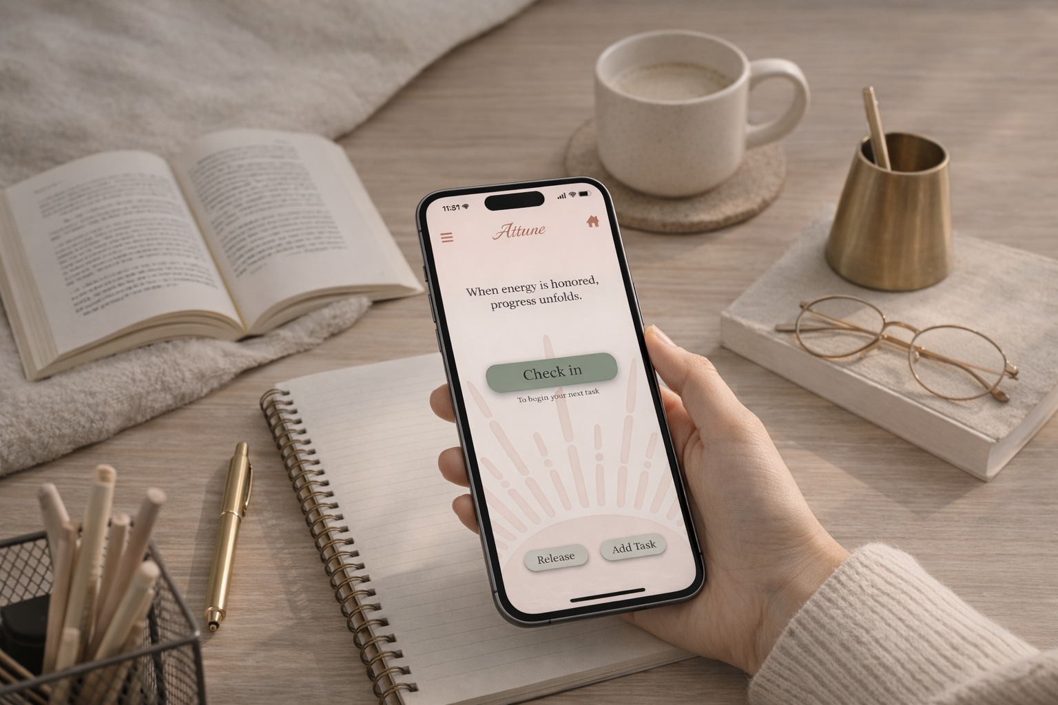 Someone holding a smartphone displaying a check-in app, with a notebook, a pen, an open book, a pair of glasses, a cup of coffee, and a gold pen holder on a wooden desk.