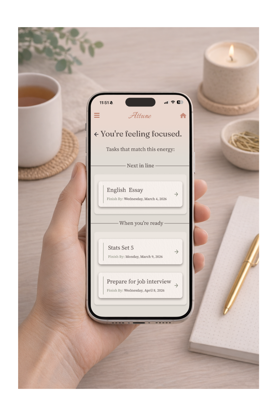 A person holding a smartphone displaying a focus tracking app with a to-do list. The app shows tasks like 'English Essay,' 'Stats Set 5,' and 'Prepare for job interview.' The workspace includes a cup of tea, a lit candle, a small bowl, a notebook, and a gold pen on a light wooden surface.