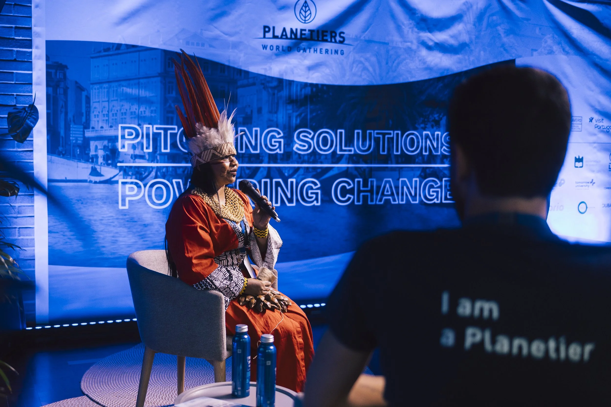 Woman in traditional Brazilian Indigenous attire speaking into a microphone, seated on a chair at an event with a blue backdrop that reads 'PITCHING SOLUTIONS, POWERING CHANGE'.
