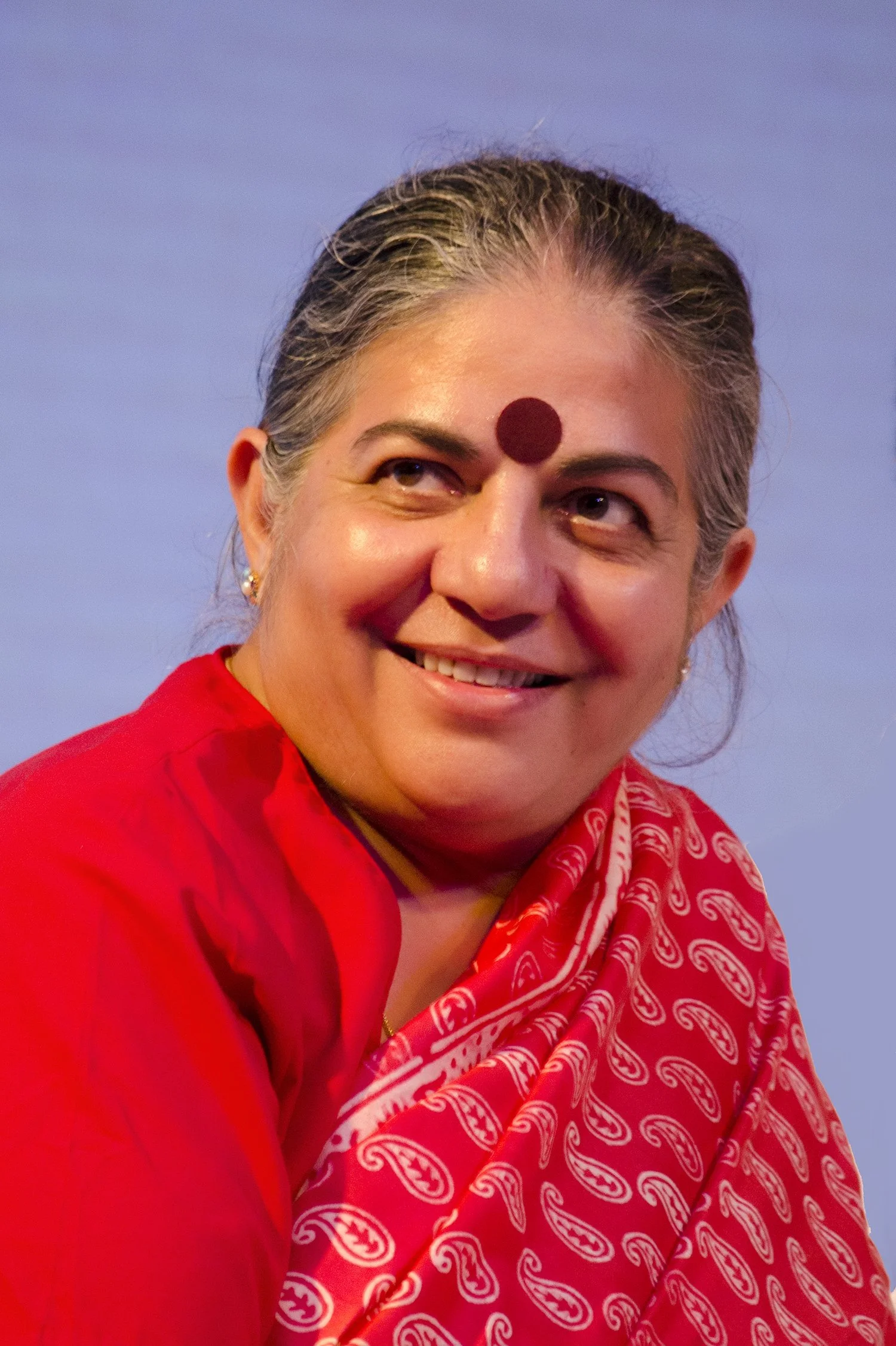 A woman with gray hair, smiling, wearing a red saree and a red bindi on her forehead.