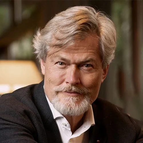 A middle-aged man with gray hair, a beard, and mustache, wearing a dark jacket and white shirt, smiling in an indoor setting with blurred background.