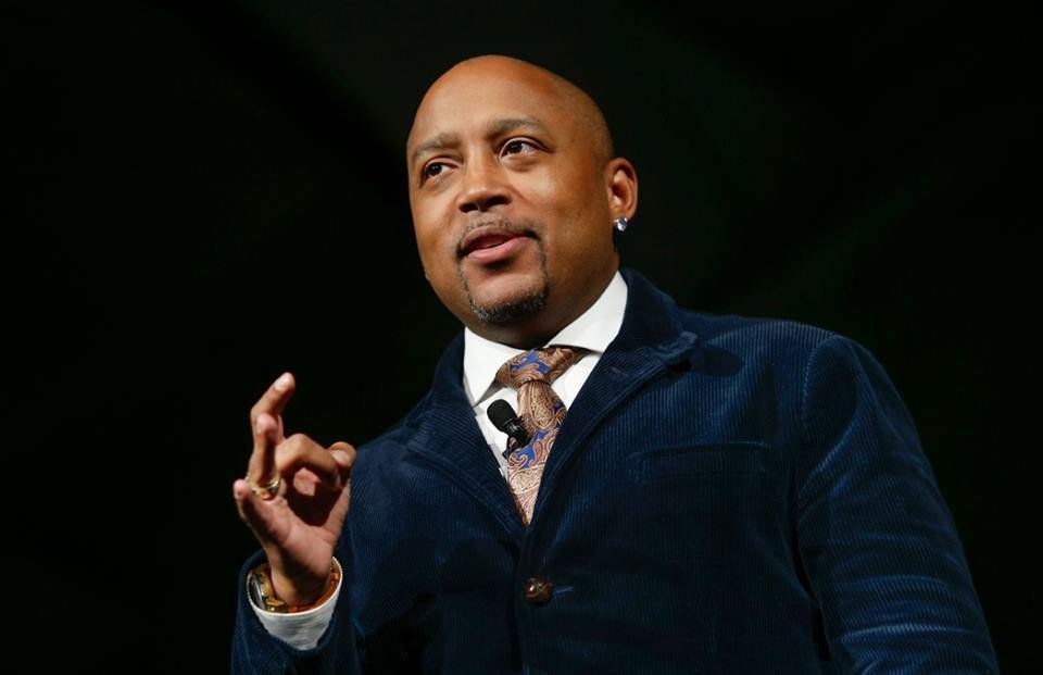 An African American man in a dark blue blazer, white shirt, and patterned tie, speaking on stage with a black background.