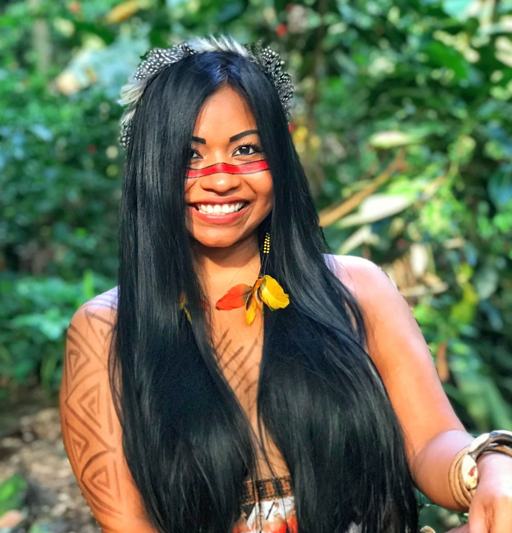 A smiling woman with long black hair, tribal face paint across her nose, wearing traditional jewelry and a feathered headpiece, standing outdoors with lush green foliage in the background.