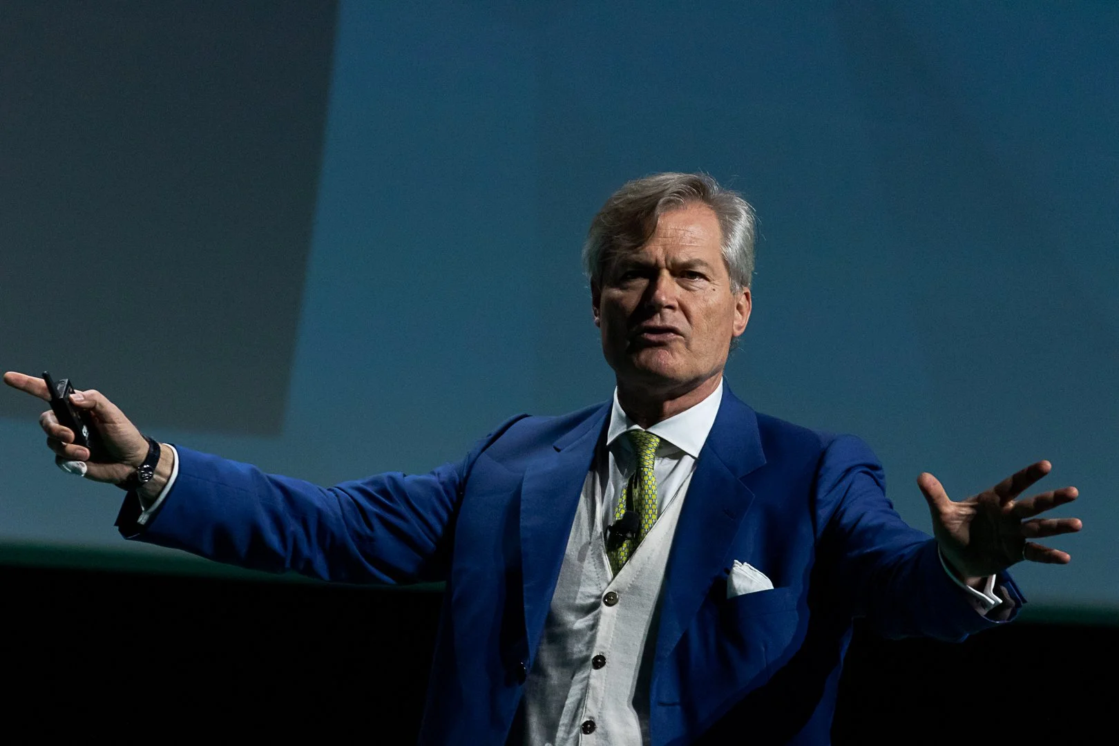 Man in a blue suit giving a presentation with his arms extended.