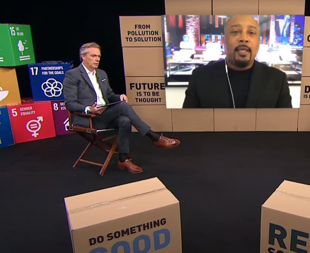 A man in a gray suit sitting on a wooden chair with a tablet, next to a large TV screen with a man speaking, and colorful boxes with slogans and the SDG icons on a black carpeted floor.