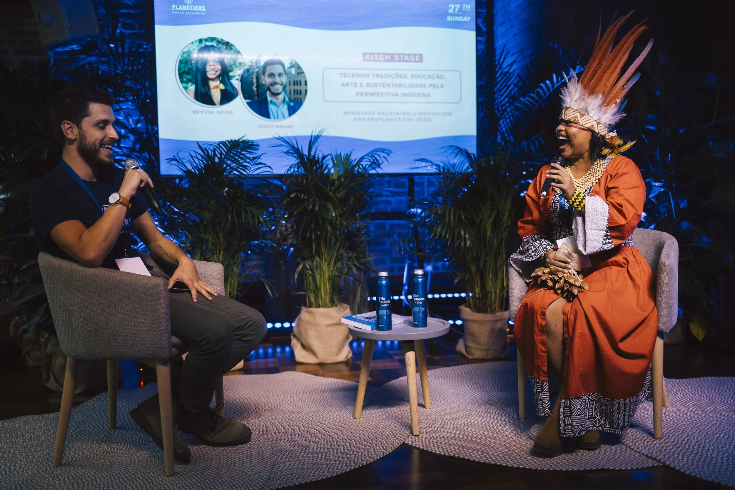 Two people seated in chairs with microphones on a stage, engaging in a conversation. The woman on the right is dressed in traditional indigenous attire, including an ornate feather headdress, an orange dress, and beaded jewelry. The man on the left is casually dressed in a black t-shirt. Behind them, a large screen displays a presentation with photos of two individuals and text in Portuguese or Spanish. There are potted plants and water bottles on a small table between them.