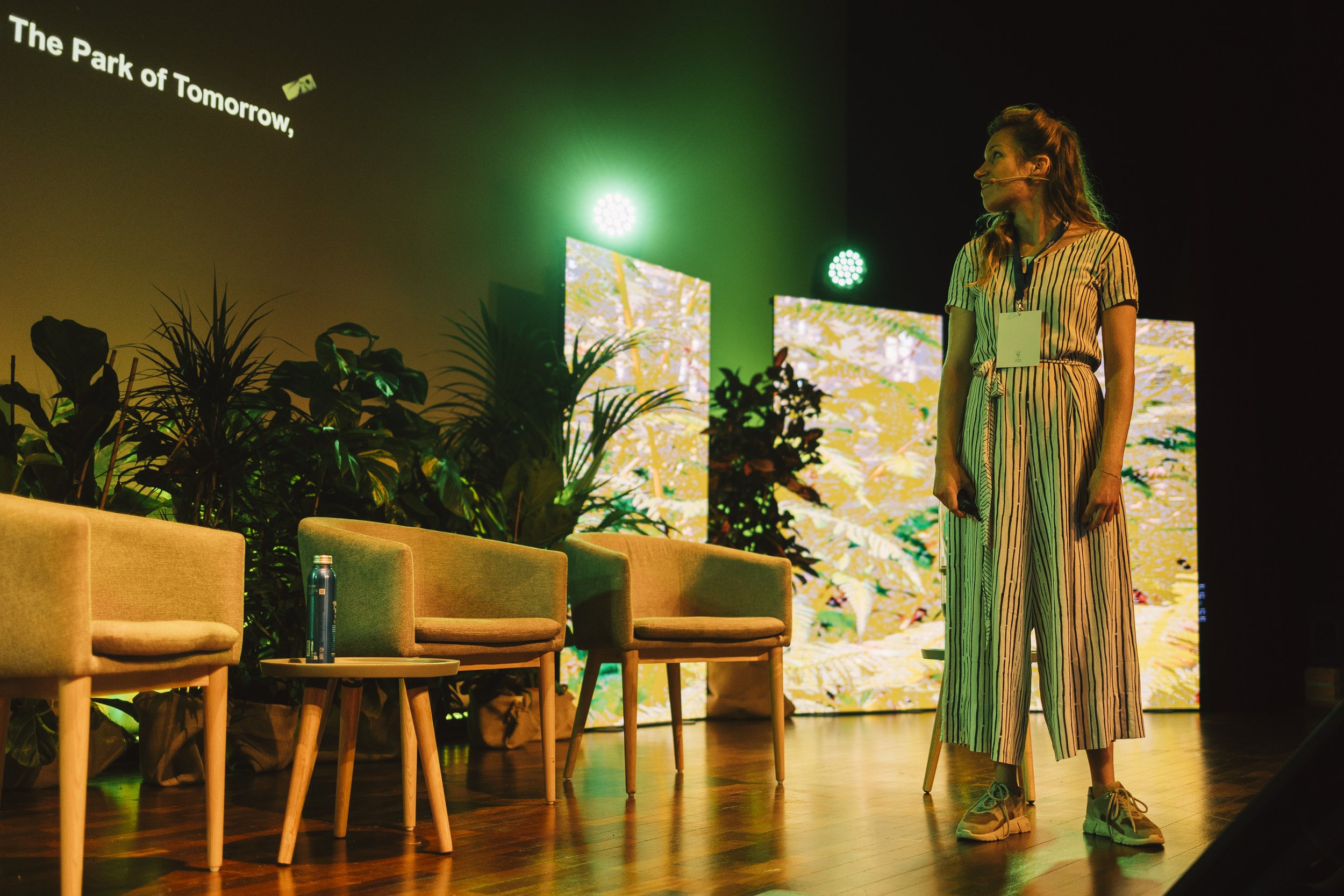 A woman in striped jumpsuit standing on a stage with green lighting and plants, behind her three green chairs and a small table with a water bottle, and a large digital screen displaying foliage.