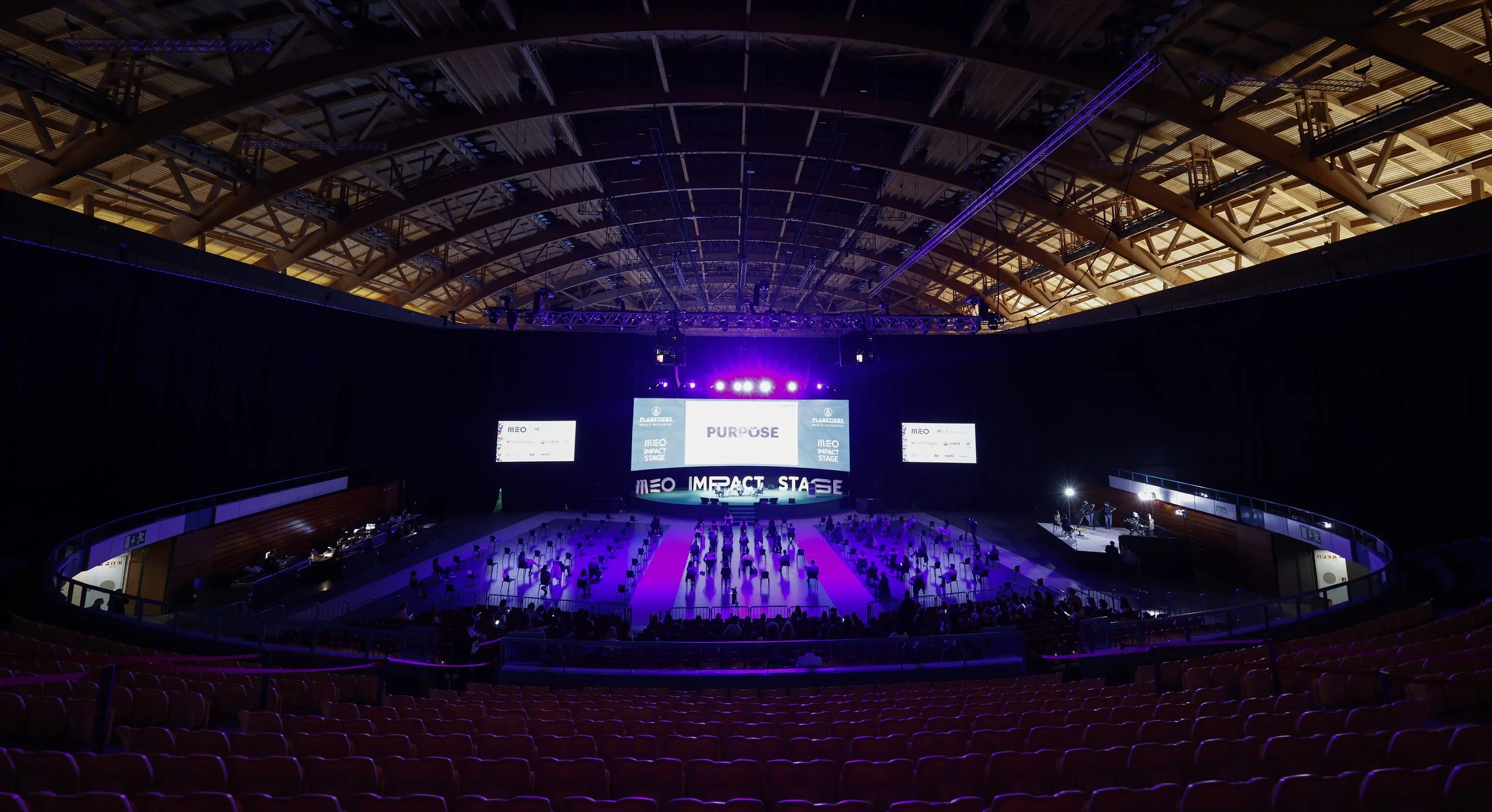 Large indoor auditorium with a stage labeled 'Impact Stage' and screen displays showing 'Purpose.' Seating arranged with social distancing spacing, purple lighting, and a mostly empty audience area.