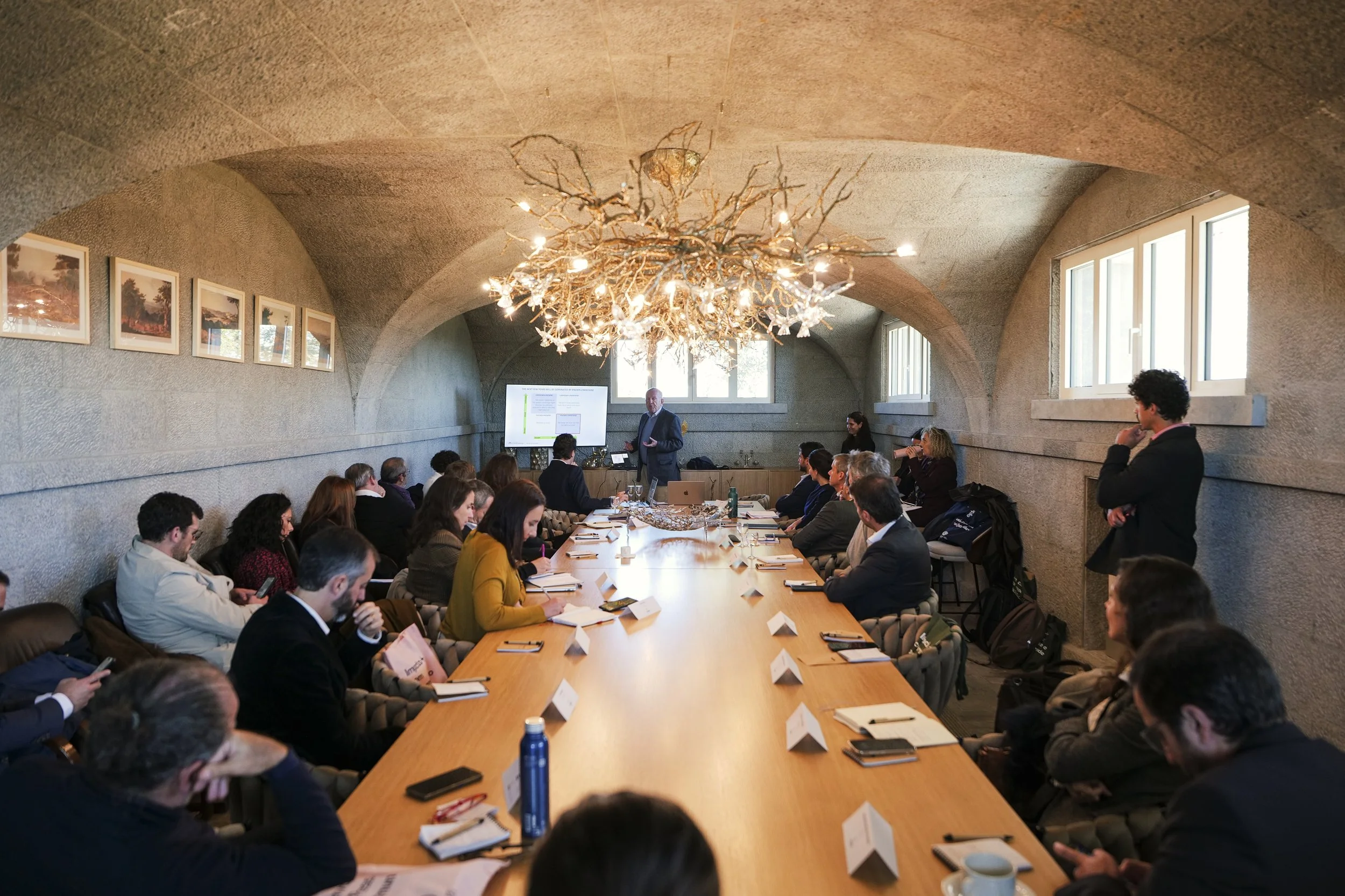 A conference room with a long wooden table surrounded by people attending a presentation. A man is standing near a screen at the front, speaking to the audience. The room has arched ceilings, large windows, and a chandelier hanging from the ceiling.
