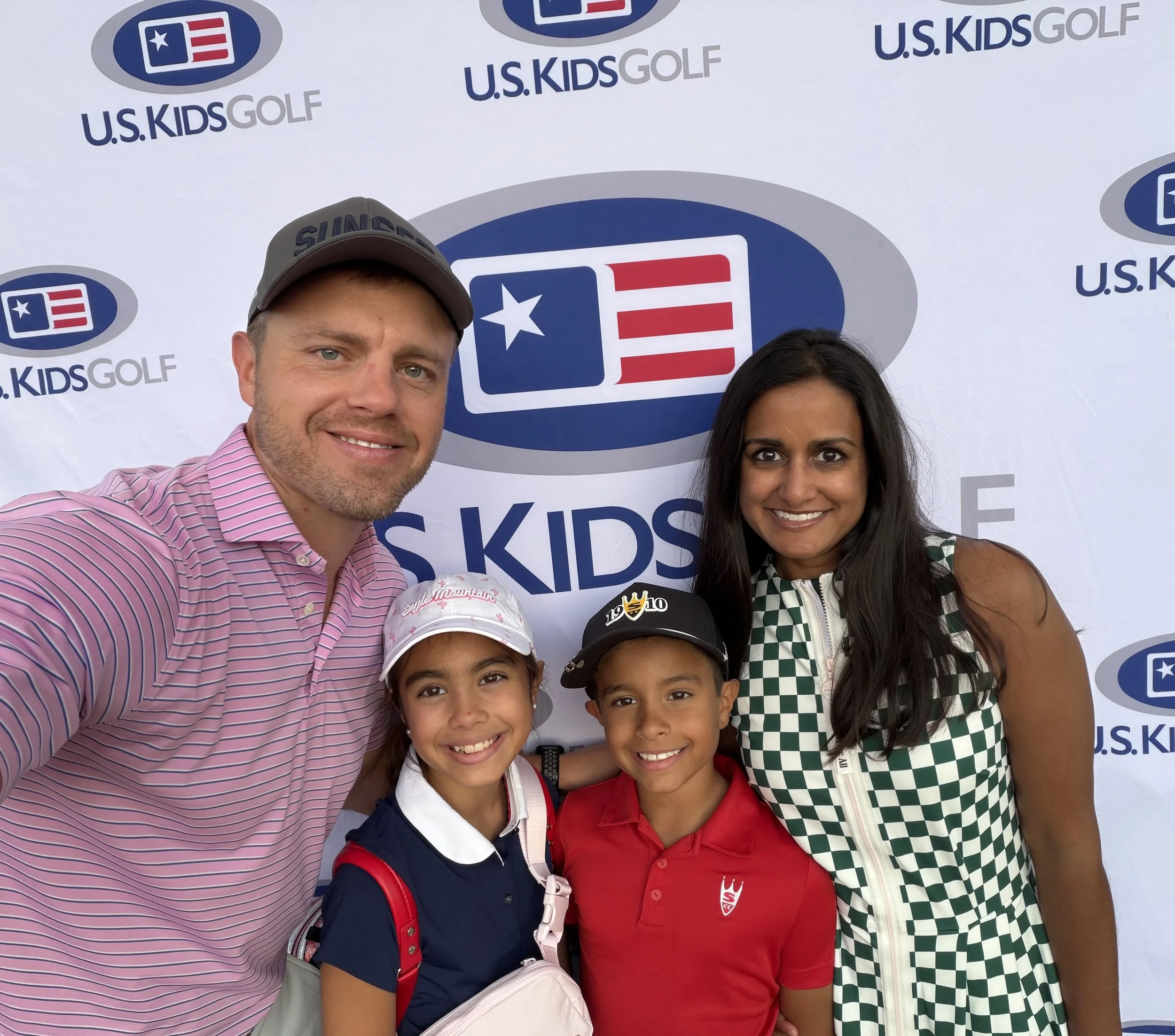 A family of four smiling at a U.S. Kids Golf event, with a backdrop displaying the U.S. Kids Golf logo. The father is wearing a pink striped shirt and a gray cap, the mother is in a green and white checkered sleeveless dress, and the children are a girl in a navy polo and white hat, and a boy in a red polo with a black cap.