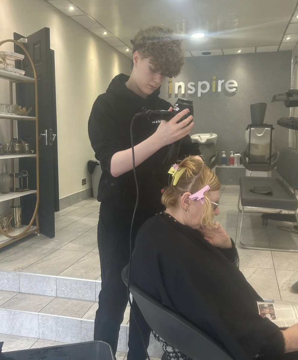 A hairstylist is blow-drying a woman's hair in a salon. The woman is seated and has her hair clipped and wrapped in yellow and pink hair clips, and she is reading a magazine. The salon has a modern interior with shelves, chairs, and a sign on the wal