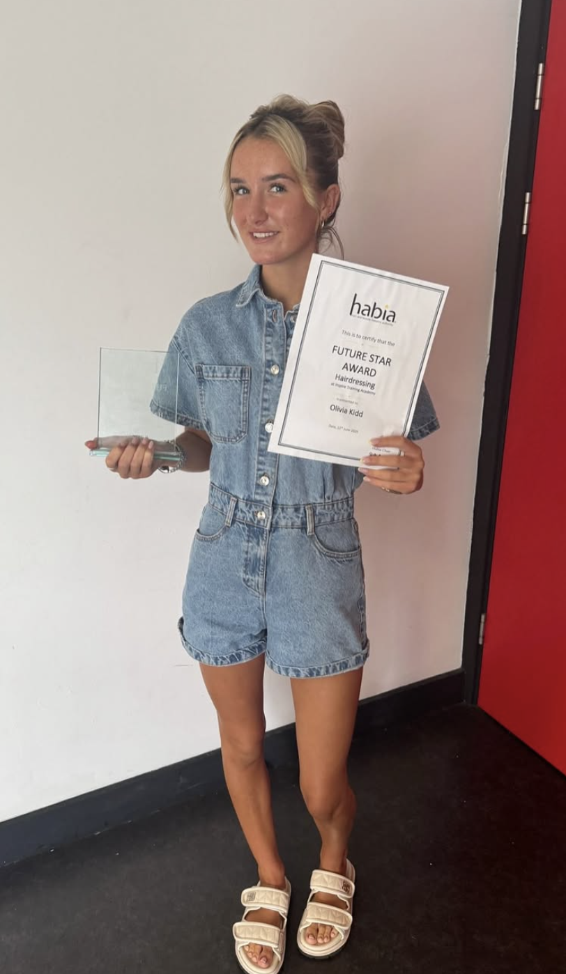 Young woman in denim outfit holding a plaque and a glass award, standing against a plain wall near a red door, celebrating an award achievement in hairdressing.