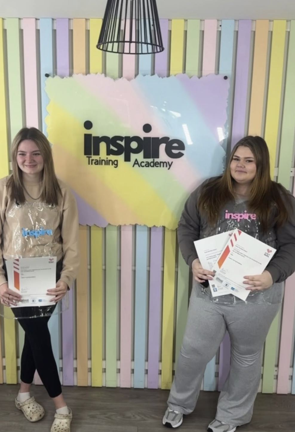 Two young women standing in front of a colorful wall with a sign that reads 'Inspire Training Academy.' Each woman is holding certificates and smiling, indicating they attended a training or educational event.