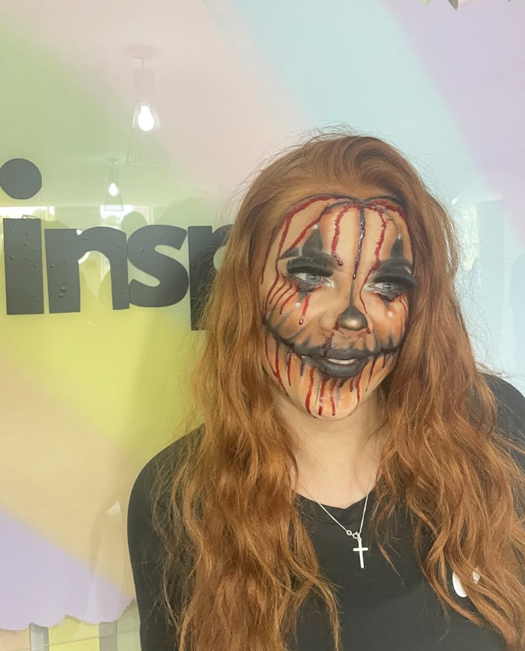Woman with Halloween makeup that resembles a creepy clown or doll face with blood streaming from the eyes and mouth, silver metallic nose, and long red hair standing in front of a sign with partially visible black text on a yellow background.