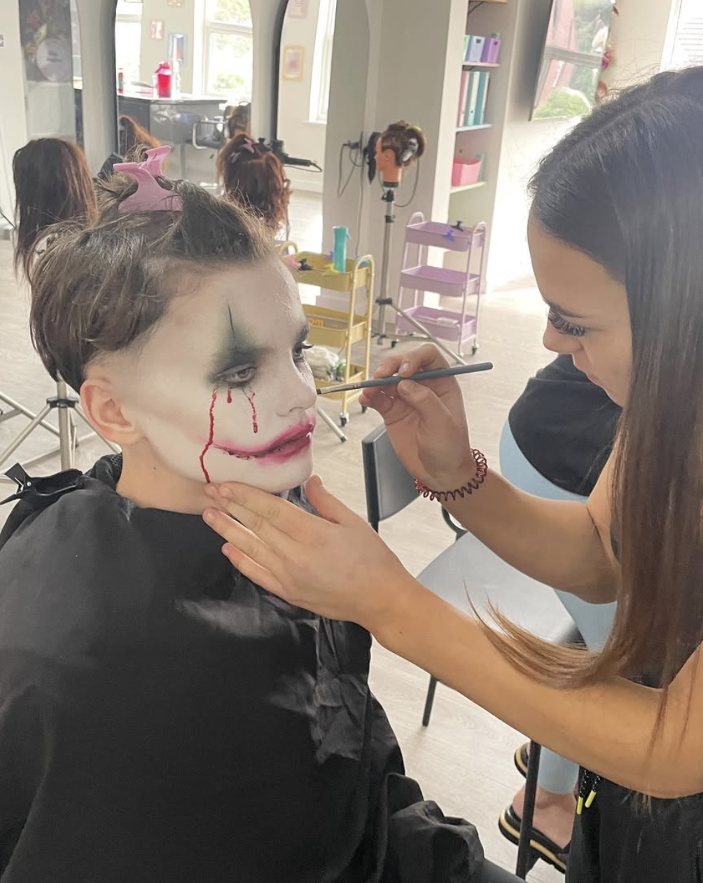 A makeup artist is applying face makeup to a young person in a clown or Joker costume, with face paint resembling a scary clown, including green and white face paint, red tears, and a painted smile.