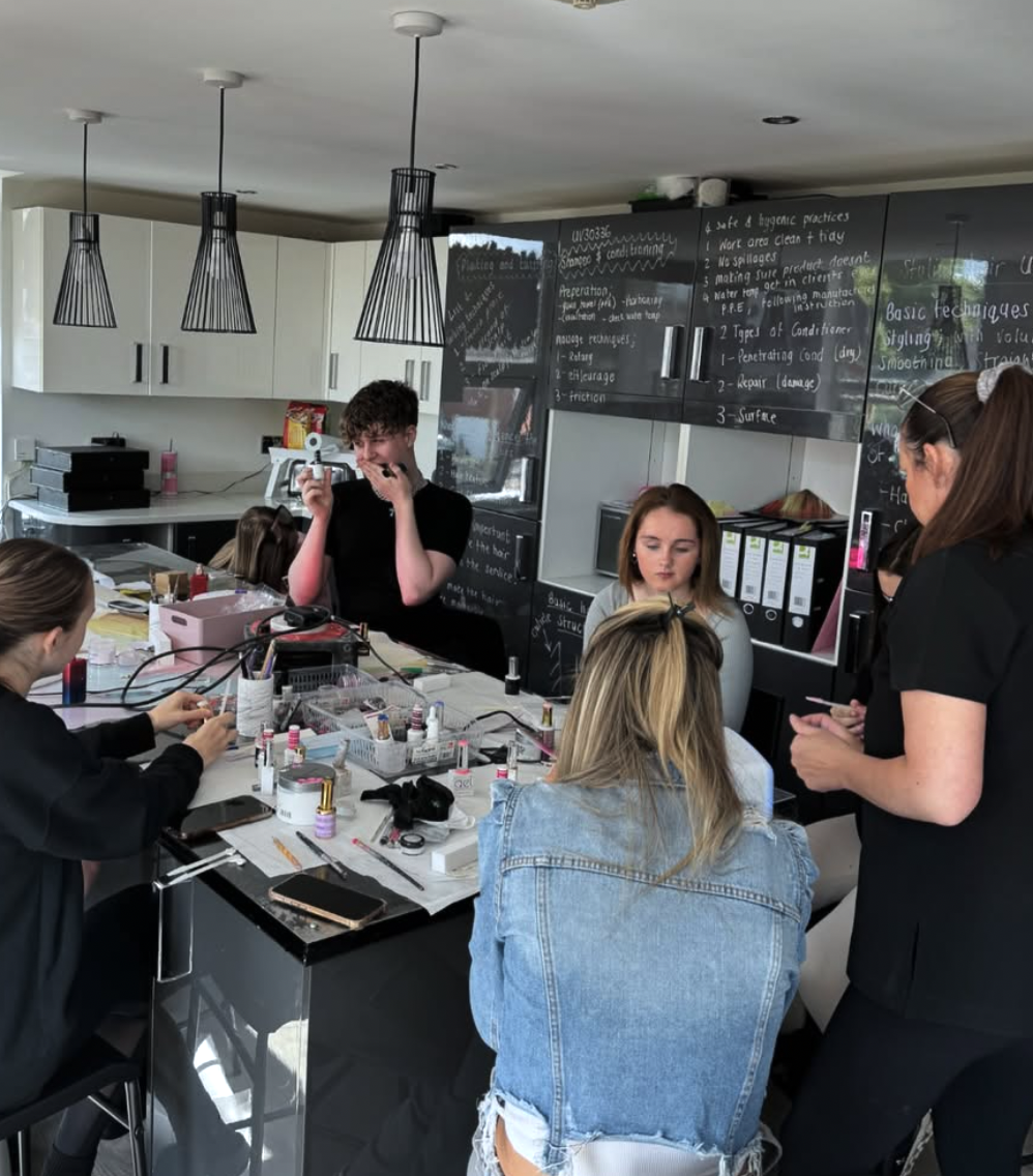 A group of women and a young man are gathered around a table in a classroom or workshop setting that appears to be a nail or beauty salon training session. The table is cluttered with nail polish bottles, tools, and other beauty supplies. The backgro