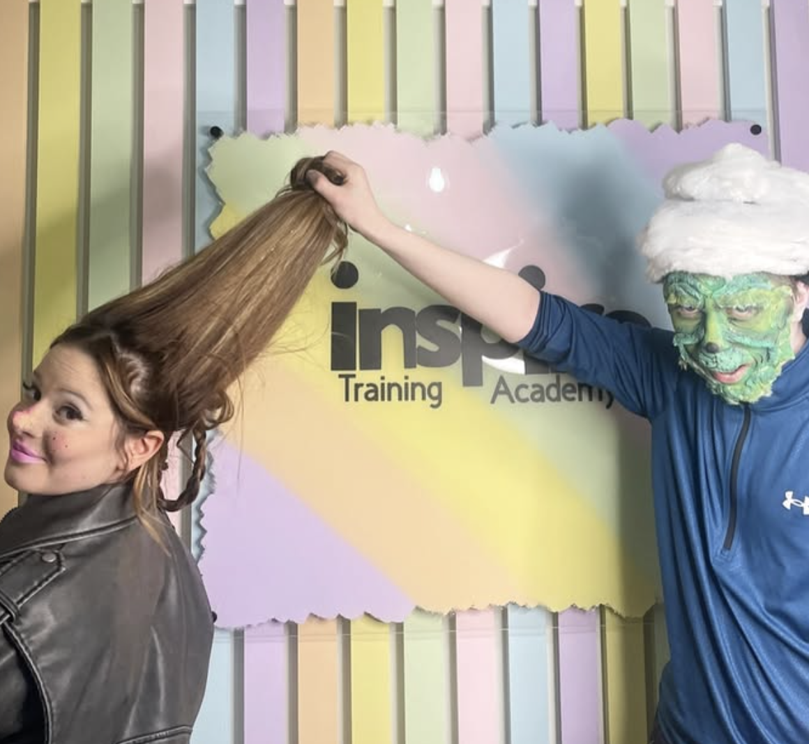 Person with face painted as a green monster wearing a Santa hat, holding a girl's long hair that is styled next to a sign for Inspire Training Academy.