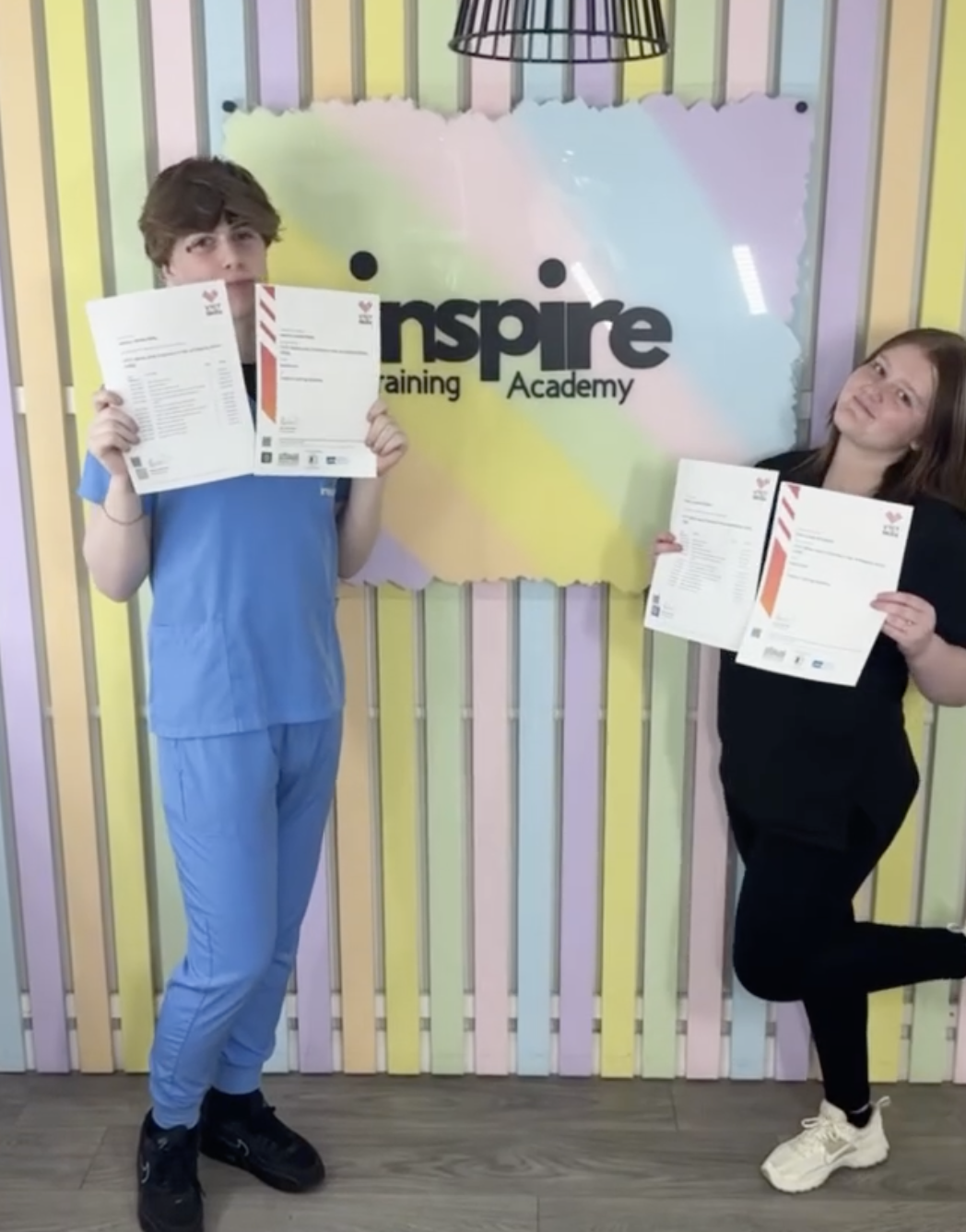 Two children standing in front of a colorful wall with 'Inspire Training Academy' written on it, each holding a paper, smiling.