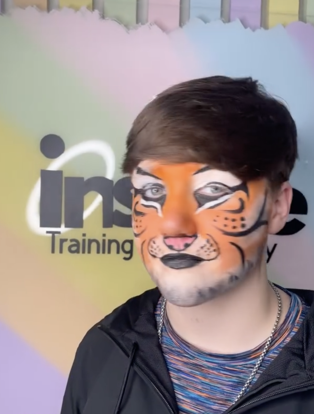 Person with tiger face paint standing in front of a colorful background with partially visible words 'Inspire Training'.