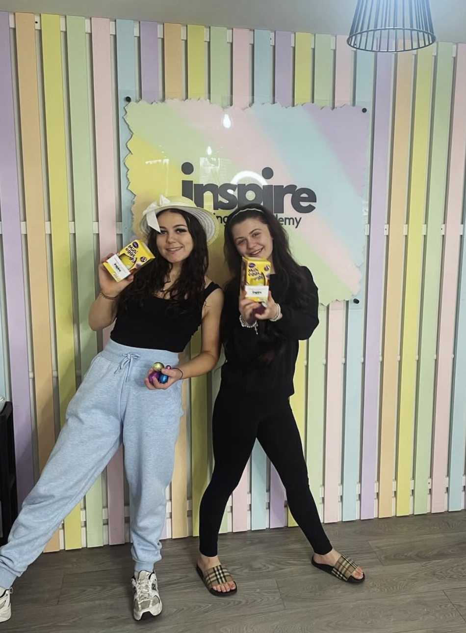 Two young women standing in front of a pastel-colored wooden wall at Inspaire Learning Academy, holding boxes of Crayola Crayons and smiling at the camera. One is wearing a black tank top, light gray sweatpants, white sneakers, and a white hat, holdi