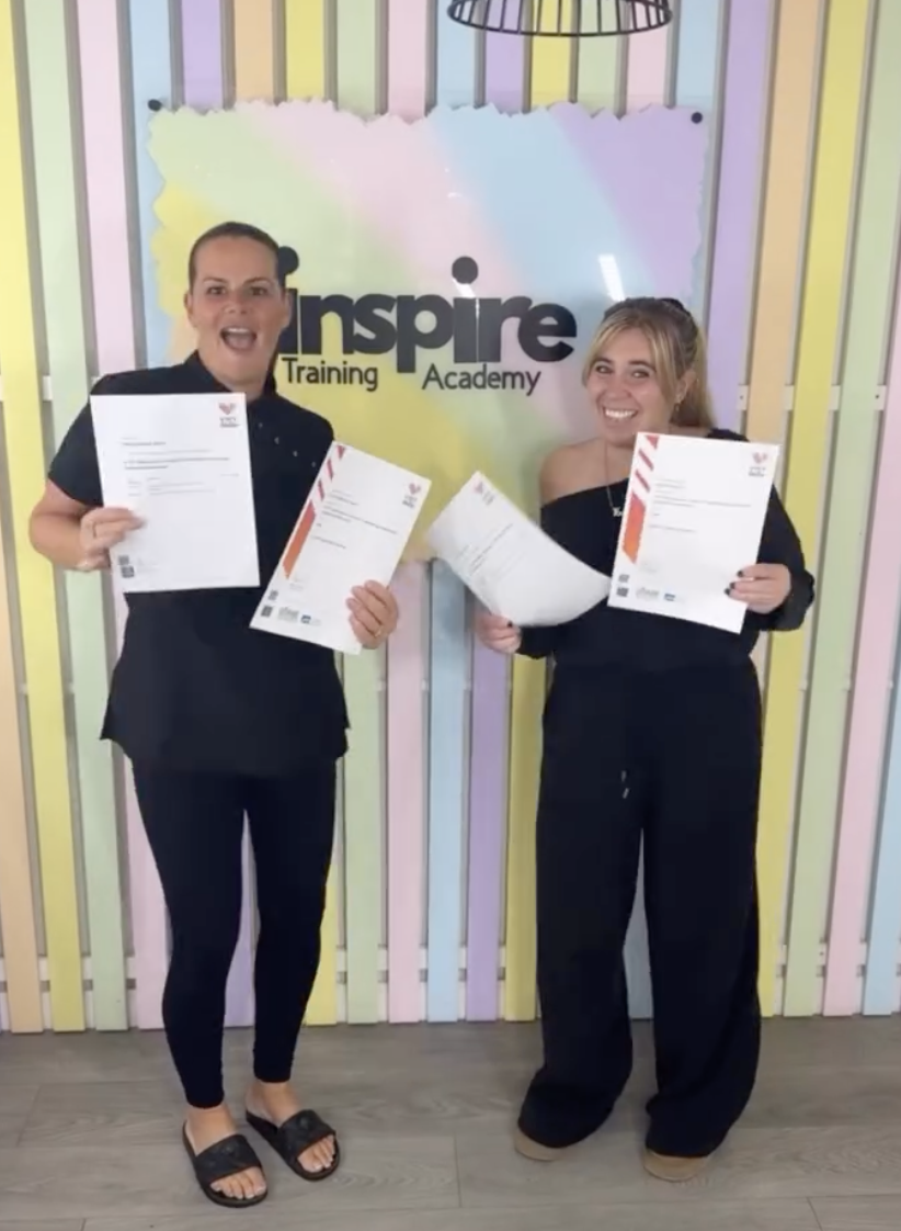 Two women standing in front of a colorful wall with the words 'Inspire Training Academy'. They are smiling and holding certificates, indicating they have completed training or courses.