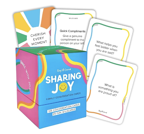 A colorful box labeled "Sharing Joy Family Conversation Cards" contains 125 conversation cards and 25 fun wildcards. Several cards are fanned out, showing prompts such as "Cherish every moment," "Quick compliments," "What helps you feel better when you are sad?", and "What is something you are proud of?".
